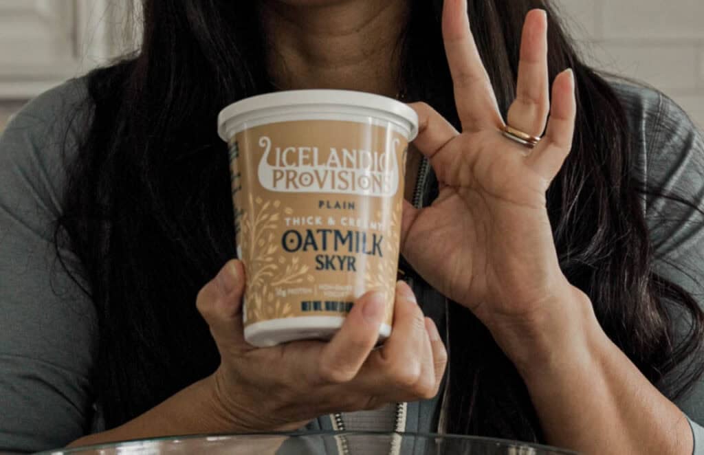 A person with long dark hair holds a container of Icelandic Provisions plain oatmilk skyr. Their left hand is making an OK gesture, and they are wearing a gold ring.